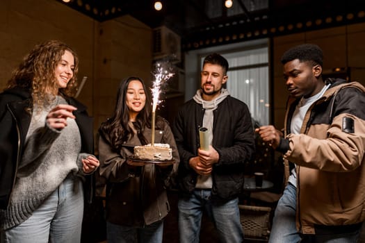A group of four friends joyfully celebrate a birthday inside a cozy venue. One person holds a cake topped with sparklers as the others cheer and enjoy the moment together.