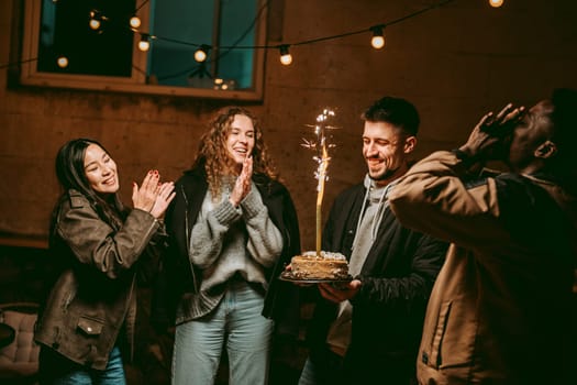 Four friends joyfully celebrate with a cake featuring sparklers on a cool evening. Laughter fills the air under warm string lights, creating a festive mood.