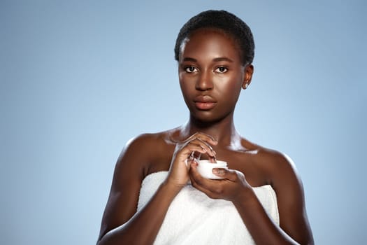 A woman stands with a towel wrapped around her, applying cream to her skin. The soft blue backdrop enhances the focus on her natural beauty and skincare routine, highlighting self-care.