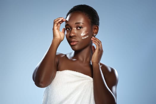 A woman with short hair lightly applies a cream to her face while wearing a towel. She appears focused and confident in a well-lit studio environment, showcasing a beauty routine.