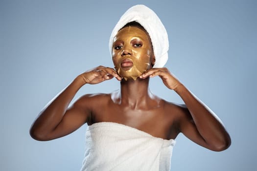 A woman stands confidently, wearing a gold facial mask and a towel on her head. She is preparing for a skincare routine in a bright, calming environment, focusing on her beauty regimen.