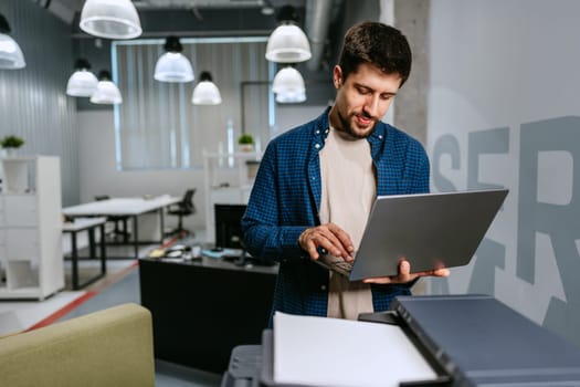 A man is standing in a contemporary office environment, focused on his laptop while preparing to print documents. The workspace features modern furniture and bright lighting.