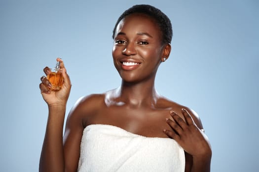 A woman with short hair is smiling while holding a perfume bottle in a bright studio. She is wearing a white towel and exudes confidence and joy. The soft blue backdrop enhances the mood.