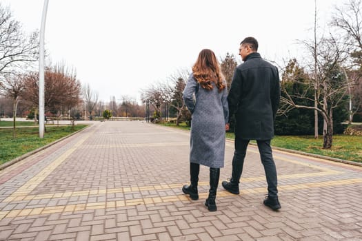 A couple strolls hand in hand along a tranquil park pathway lined with trees. The air is cool, and the sky is overcast, creating a calm atmosphere for their walk.