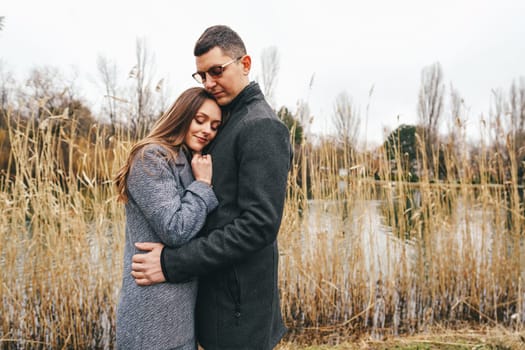 A couple stands closely together by a quiet body of water, surrounded by tall reeds. The woman rests her head on the man's shoulder, both wearing warm coats as they enjoy a calm autumn day.