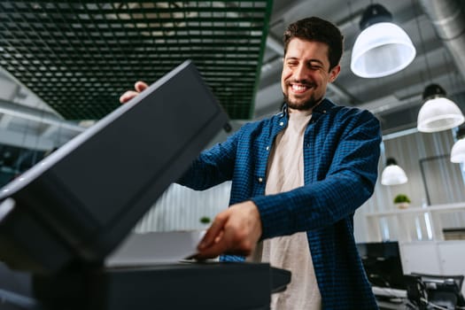 A man smiles while interacting with a printer in a contemporary office space filled with overhead lights and computers. He appears focused on his task.
