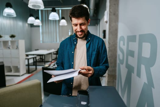 A young man stands in a contemporary office, smiling as he reviews printed papers from a printer. Natural light fills the workspace, creating an inviting atmosphere for productivity.