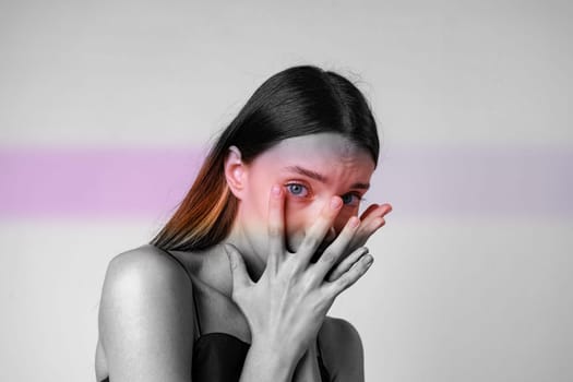 A woman stands in a minimalist setting, daylight illuminating her features. She expresses deep emotion with her hands covering her face, revealing striking blue eyes.
