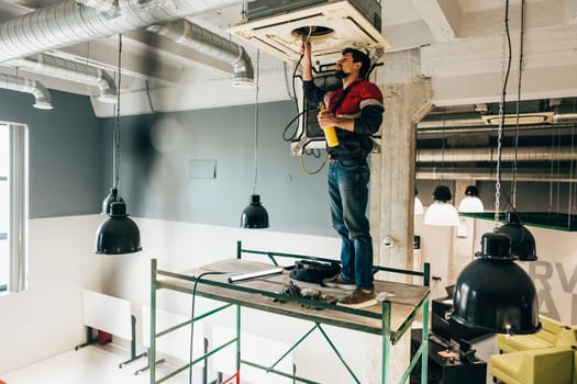 A technician is reaching up to repair a ventilation system in a commercial building. The setting features exposed ductwork and an industrial aesthetic, illuminated by natural light.