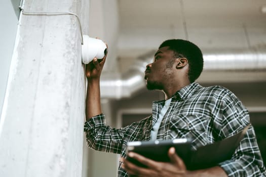 A technician adjusts a security camera mounted on a concrete pillar in a contemporary office setting. He holds a tablet while ensuring proper alignment and functionality.