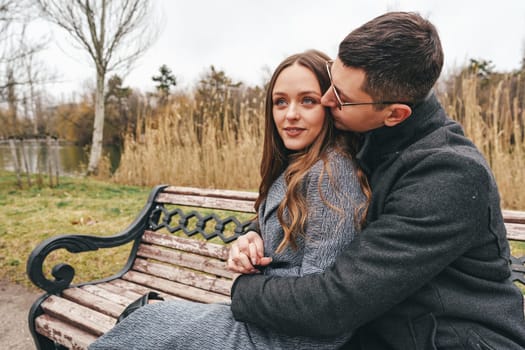 A young couple sits closely on a park bench, sharing a warm moment. They are surrounded by tall grass and trees in a serene outdoor setting during autumn.