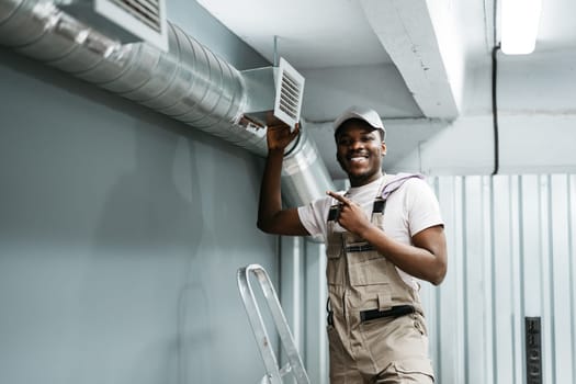 A professional worker wearing a cap and overalls stands next to a newly installed air vent in a workshop. The person is smiling and pointing, showcasing expertise.