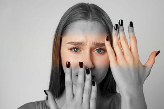 A young woman shows a variety of emotions with her hands partially concealing her face. Her nails are painted black, and her striking blue eyes contrast with the muted background.