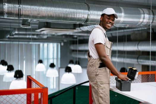 A young male worker smiles while using a tool on a table in a contemporary office environment filled with pendant lights. Bright ambiance enhances his cheerful demeanor.