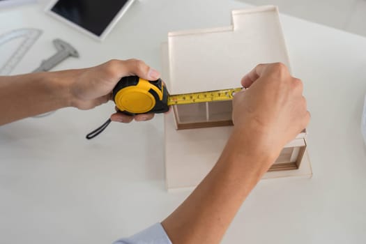 Hands carefully measuring a model house with a tape measure, showcasing attention to detail in architectural design.