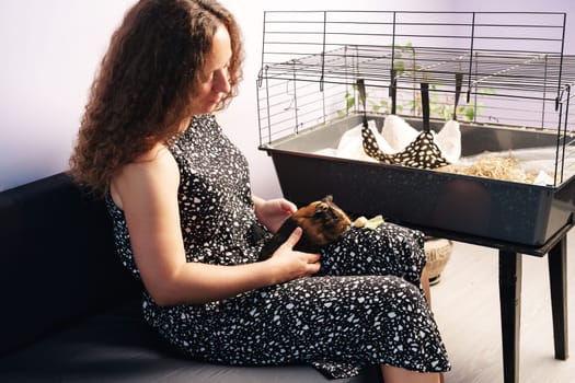 A woman with curly hair is gently holding a guinea pig while sitting on a couch. A cage is positioned nearby, filled with bedding and greenery, creating a warm atmosphere.