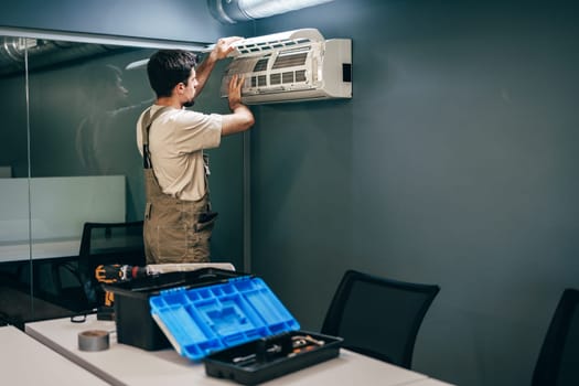 A skilled technician adjusts an air conditioning unit mounted on a wall in a contemporary office space. Tools and equipment are visible on the table nearby.
