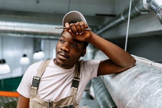 A young man stands with his hand on his forehead, wearing a cap and overalls. He appears thoughtful in a well-lit industrial workspace with metal ductwork overhead.