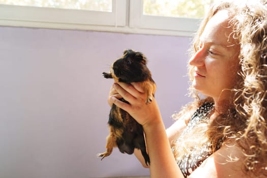 A woman gently holds a guinea pig with a smile in a bright, sunlit room. The atmosphere is warm and inviting, showcasing their special connection.