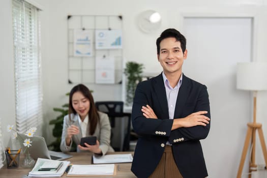 A confident businessman stands with arms crossed in a bright office, showcasing professionalism and collaboration.