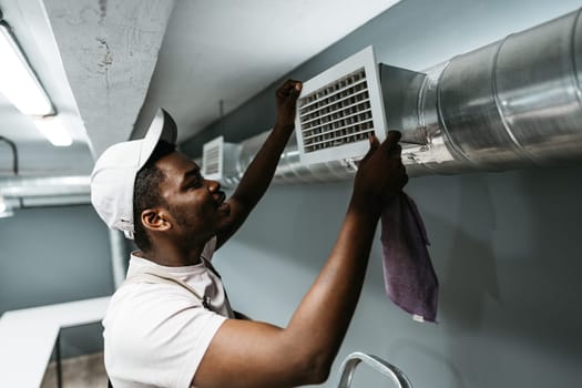 A maintenance worker is carefully adjusting an air vent in a stylish basement space. The worker wears a cap and uses a cloth for cleaning while focused on the task.