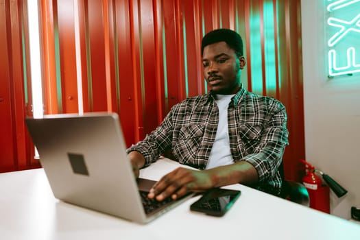 A focused young man is seated at a white table, using a laptop in a stylish workspace with red walls and green neon lights. His smartphone rests beside him as he engages in work.