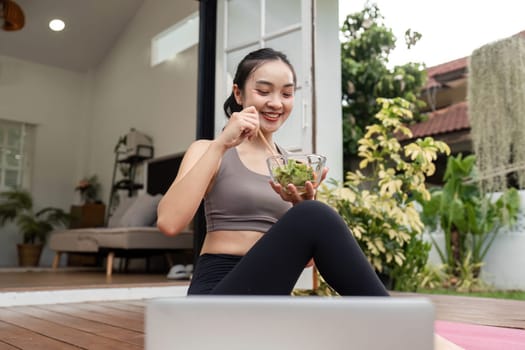 A young woman smiles while eating a fresh salad on her yoga mat, promoting healthy eating and lifestyle.