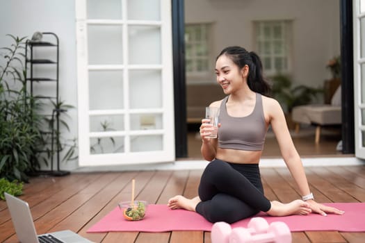 A fit woman sits on a yoga mat, drinking water and promoting healthy living and fitness while enjoying her home space.