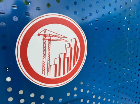 Red construction themed warning sign with cranes and skyscrapers printed on perforated blue metal fencing, symbolizing active development zone and modern urban growth initiatives