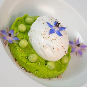 A fresh pea puree is beautifully plated, featuring a poached egg at the center and adorned with borage flowers. The dish showcases vibrant colors and textures, perfect for any spring menu.