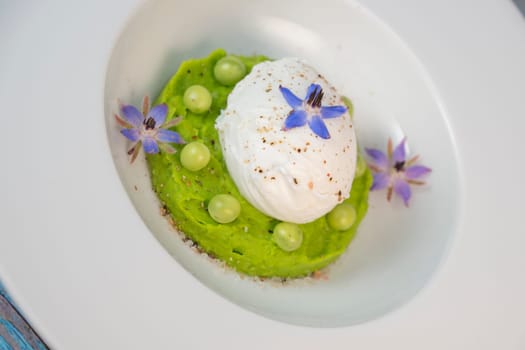A fresh pea puree is beautifully plated, featuring a poached egg at the center and adorned with borage flowers. The dish showcases vibrant colors and textures, perfect for any spring menu.
