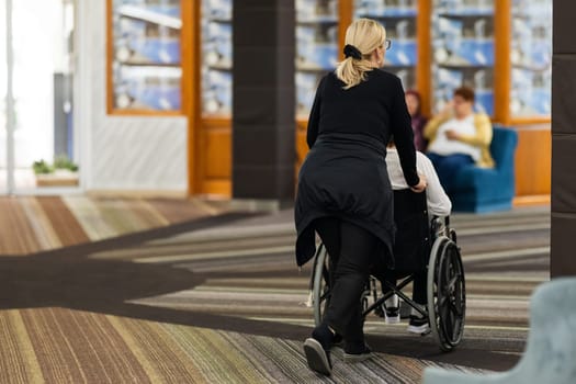 Rear view of a female caregiver pushing an elderly person in a wheelchair through a hallway.