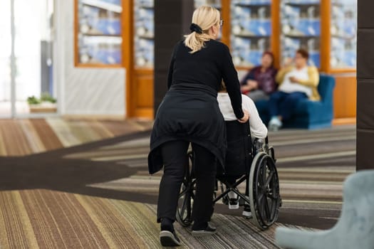 Rear view of a female caregiver pushing an elderly person in a wheelchair through a hallway.