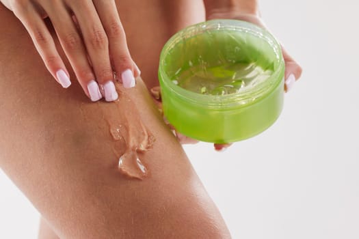 Close-up of woman applying clear gel from a green jar onto her leg, representing skincare, hydration, and body moisturizing routine on a white background.