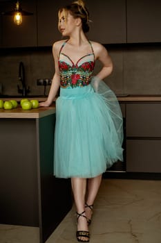 Elegant woman in turquoise skirt and corset gracefully poses in dark minimalist interior. demonstrating style, happiness and leisure. Modern kitchen, green apples on table