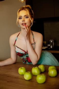 Portrait of elegant young woman in turquoise skirt and corset in kitchen interior near apples. Posing demonstrating style, happiness and leisure. Modern kitchen, green apples on table, gracefully holds hand