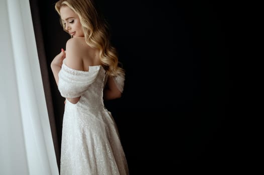 Elegant bridal gown showcased in a minimalist setting with a model in a chic pose during an indoor shoot. Blonde bride against a dark interior near the window.