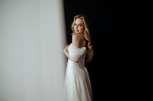 Elegant bridal gown showcased in a minimalist setting with a model in a chic pose during an indoor shoot. image of a blonde bride against a dark interior. smiles happily and looks at the camera