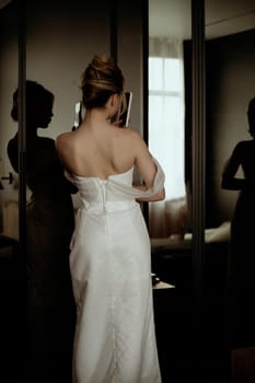 The bride looks in the mirror and takes a photo of herself on the phone. takes a selfie. view from the back of the corset of a long elegant dress.