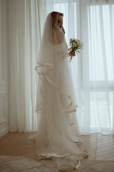 Bride with luxurious long vulya and elegant dress poses against the background of the window. huge window with silhouette of bride with bouquet of lilies. Bride waiting for groom.