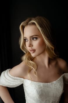 Portrait of an elegant bridal gown showcased in a minimalist setting with a model in a chic pose during an indoor shoot. Blonde bride against a dark interior near a window