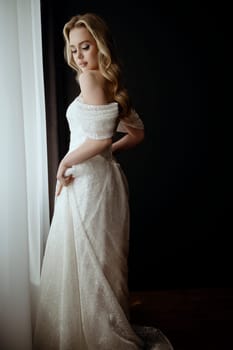 Elegant bridal gown showcased in a minimalist setting with a model in a chic pose during an indoor shoot. image of a blonde bride against a dark interior near a window.