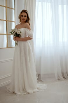 Bride with a bouquet of white flowers posing by the window. Blonde in a flowing wedding dress, radiates grace and beauty. In a light minimalist interior with a window and a mirror.