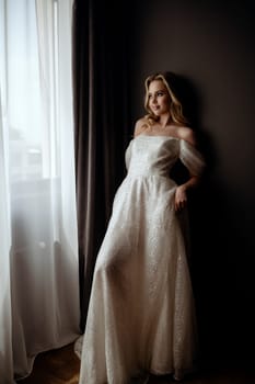 A graceful bride poses in a long, white wedding dress. In full growth against the background of a dark minimalist interior near a window. The dark background emphasizes her classic beauty