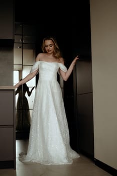 Elegant wedding dress presented in minimalist interior with model in chic pose. Blonde bride against dark interior. Bride waiting for groom