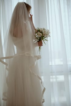 Bride with luxurious long vulya and elegant dress posing against the window. huge window with silhouette of bride with bouquet of lilies. Bride waiting for groom and wiping away tears
