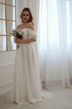 Bride, standing at full height with a bouquet of white flowers by the window. Blonde in a flowing wedding dress, radiates grace and beauty. In a light minimalist interior with a window and a mirror.