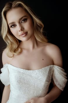 Portrait of an elegant bridal gown showcased in a minimalist setting with a model in a chic pose during an indoor shoot. Blonde bride against a dark interior near window. She is looking at the camera