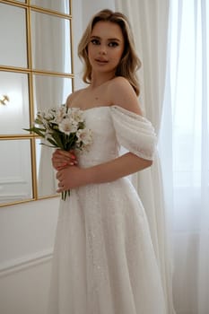 Portrait of a blonde bride with a bouquet of white flowers by the window. Blonde in a flowing wedding dress, radiates grace and beauty. In a light minimalist interior with a window and a mirror