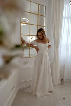 Bride, standing at full height by the window. Blonde in a flowing wedding dress, radiates grace and beauty. In a light minimalist interior with a window and a mirror.